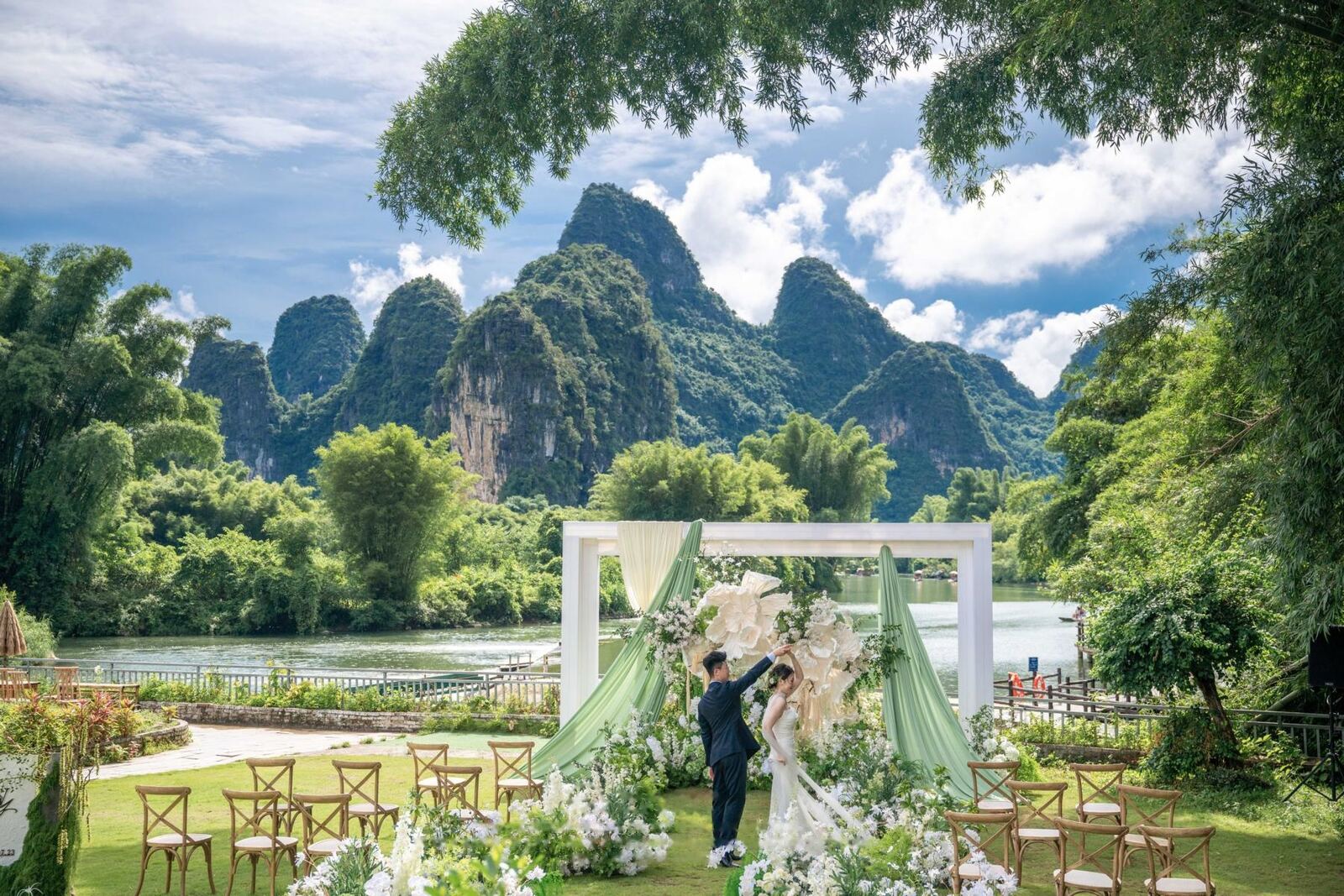 An outdoor ceremony set on a riverside lawn with karst peaks behind