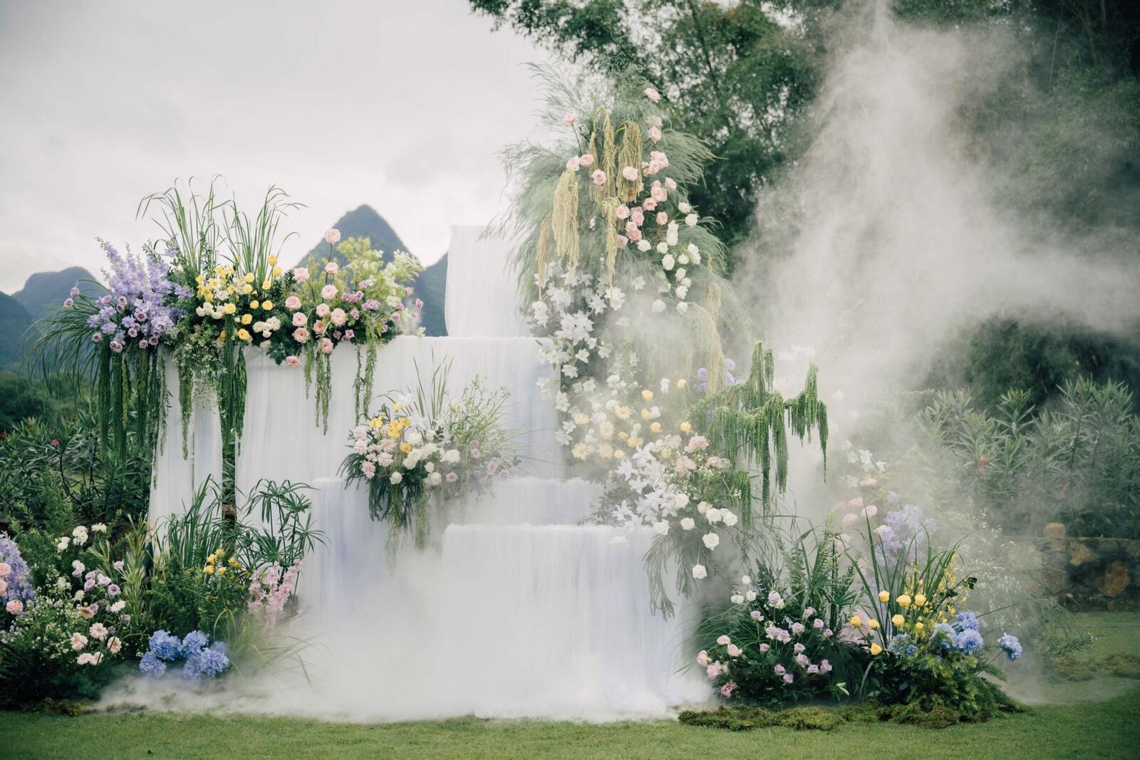 A tiered white ceremony platform with flowers and low mist on a lawn below karst hills